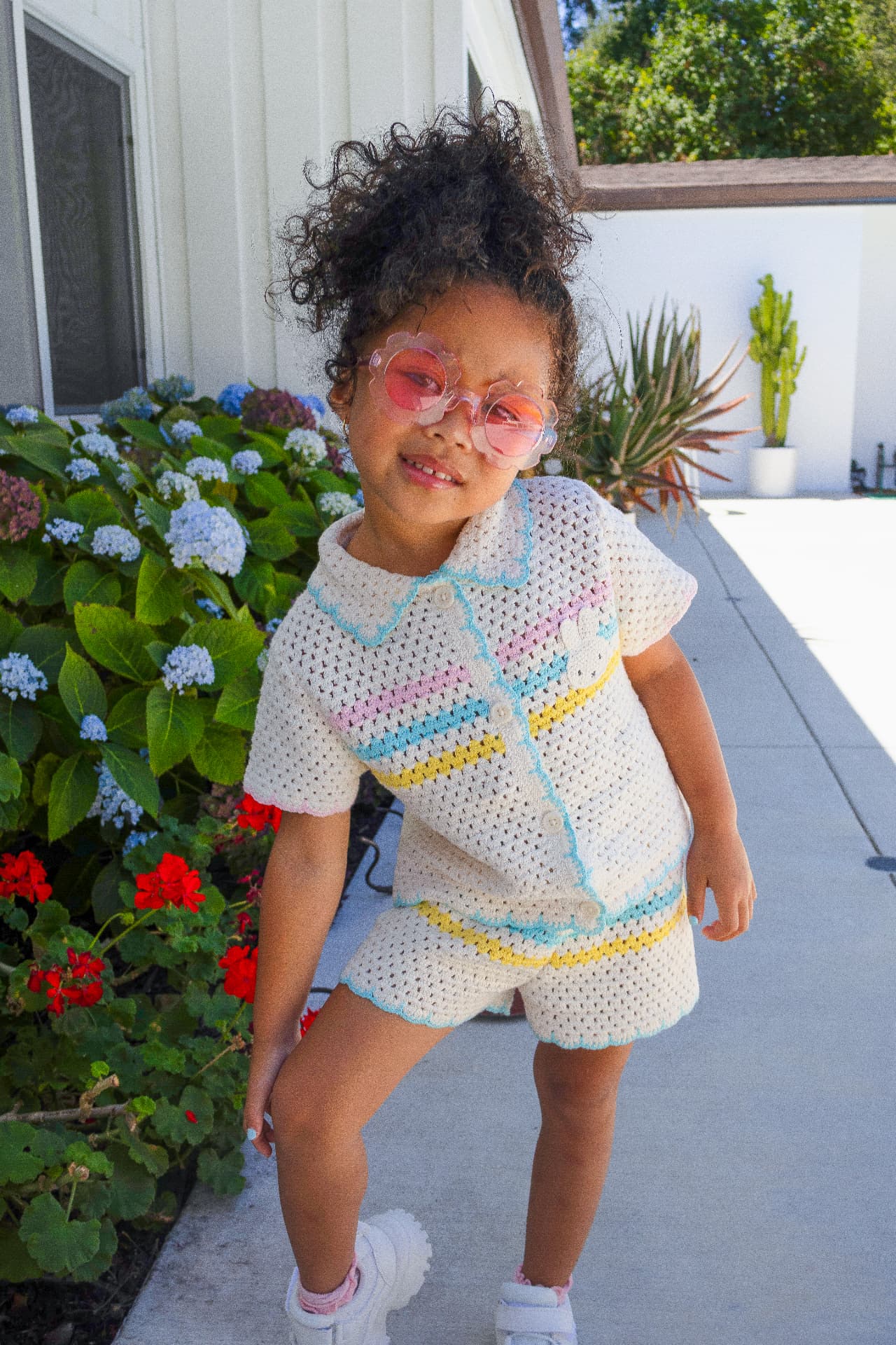 Milani posing outdoors in a white crocheted outfit with pastel stripes and pink flower sunglasses beside blooming hydrangeas