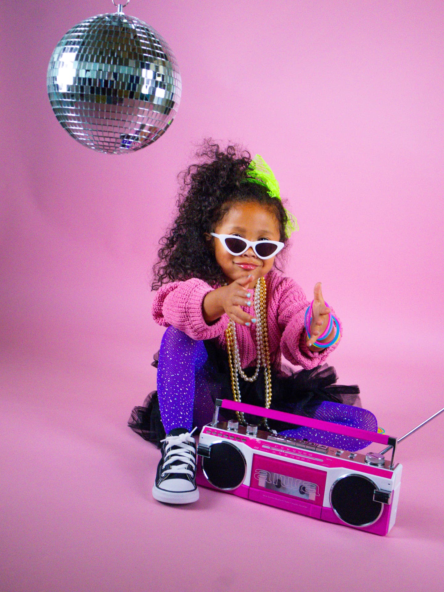 Milani sitting cross-legged in a pink knit sweater, tulle tutu, and sparkly purple leggings with a boombox, white cat-eye sunglasses and a green bow in her hair