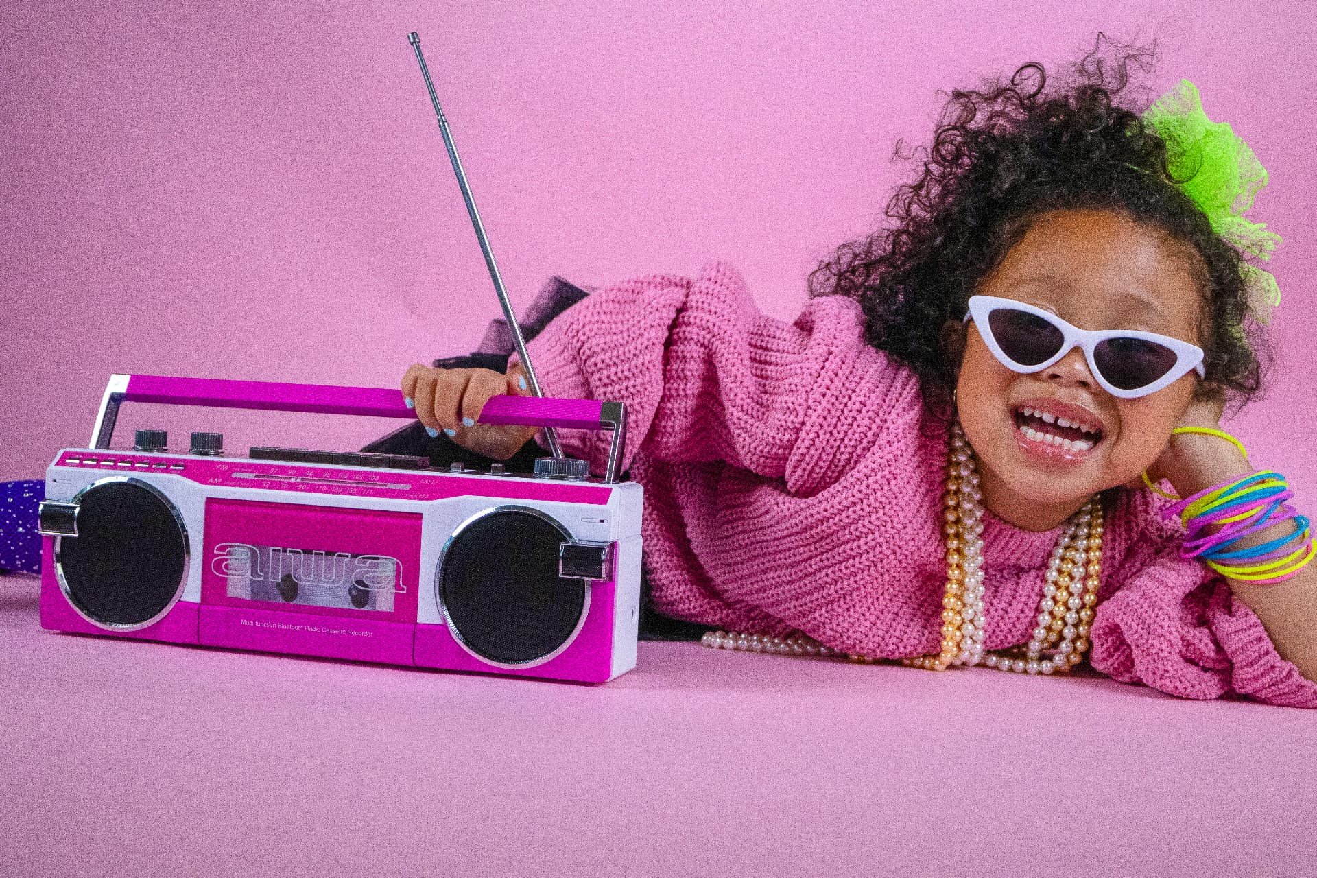 Milani lying beside a pink boombox in a pink knit sweater, white cat-eye sunglasses, and colorful bracelets, laughing