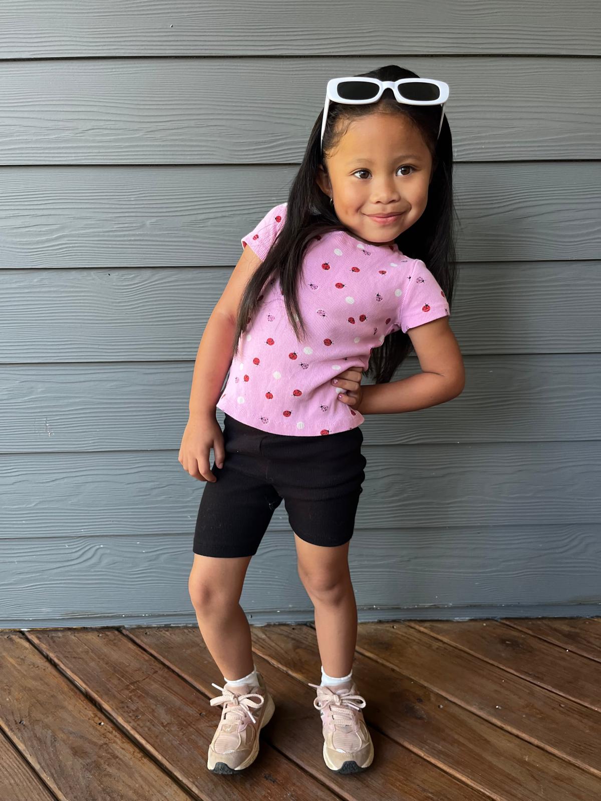 Milani in a pink ladybug tee, black shorts, and white sunglasses