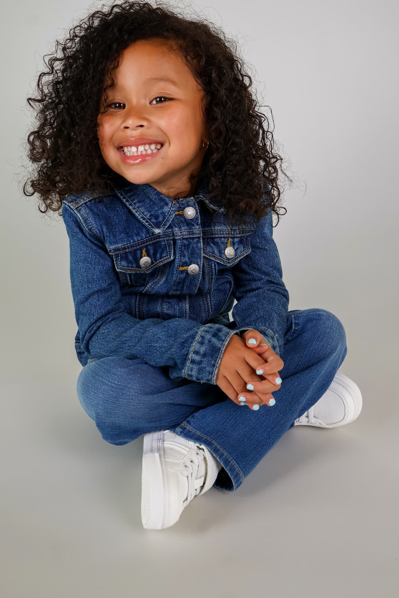Milani sitting cross-legged in a denim jacket and jeans with white sneakers, smiling in a studio portrait