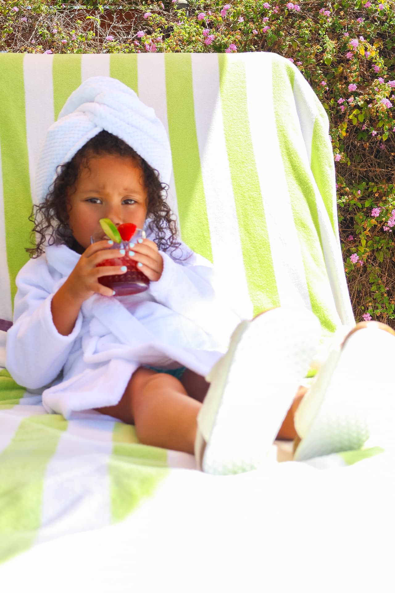 Milani relaxing in a white terry bathrobe and towel turban, sipping a red drink on a striped lounge chair in a bright garden