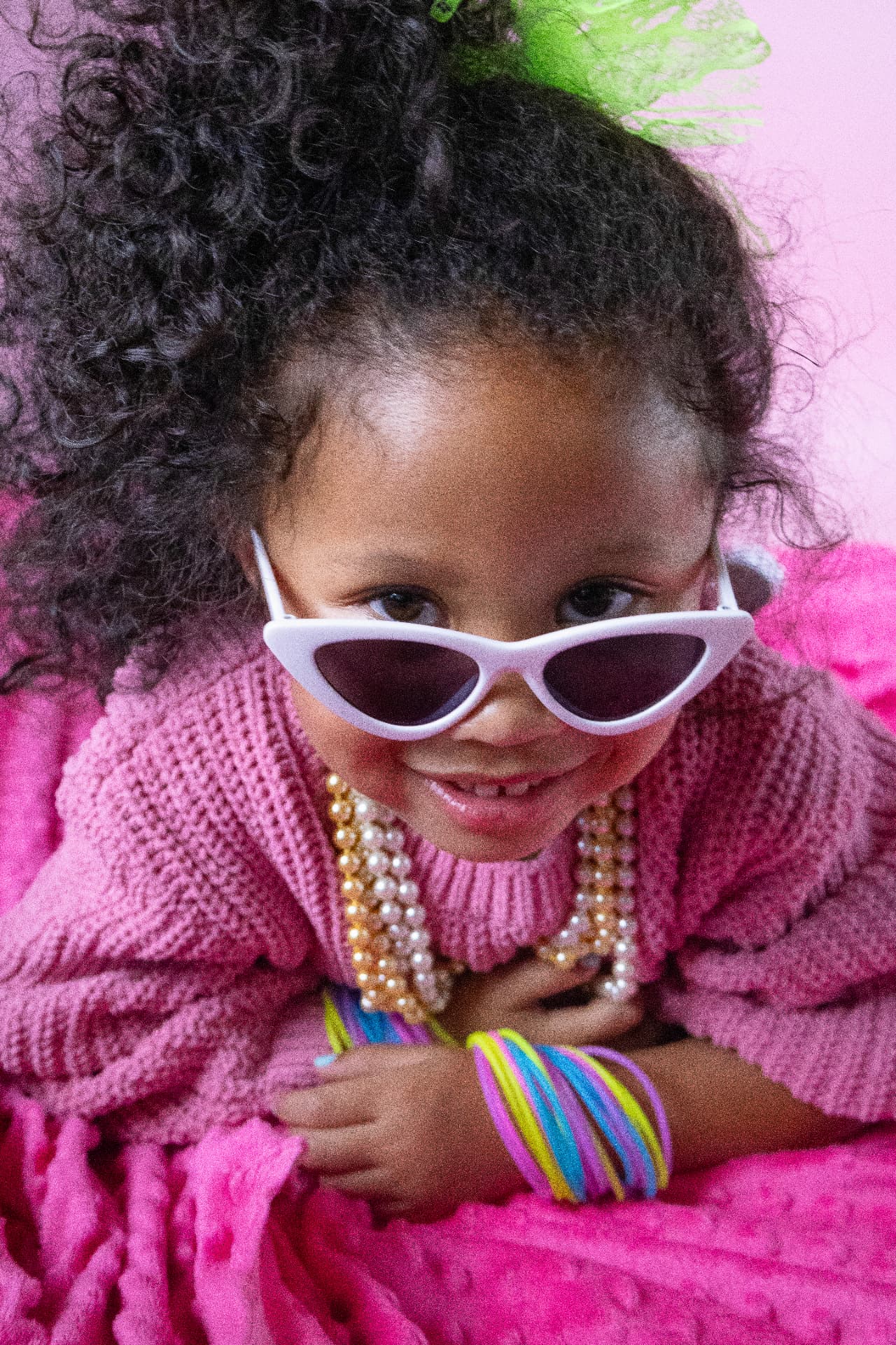 Close-up portrait of Milani in white cat-eye sunglasses, a pink sweater, pearls, and colorful bracelets