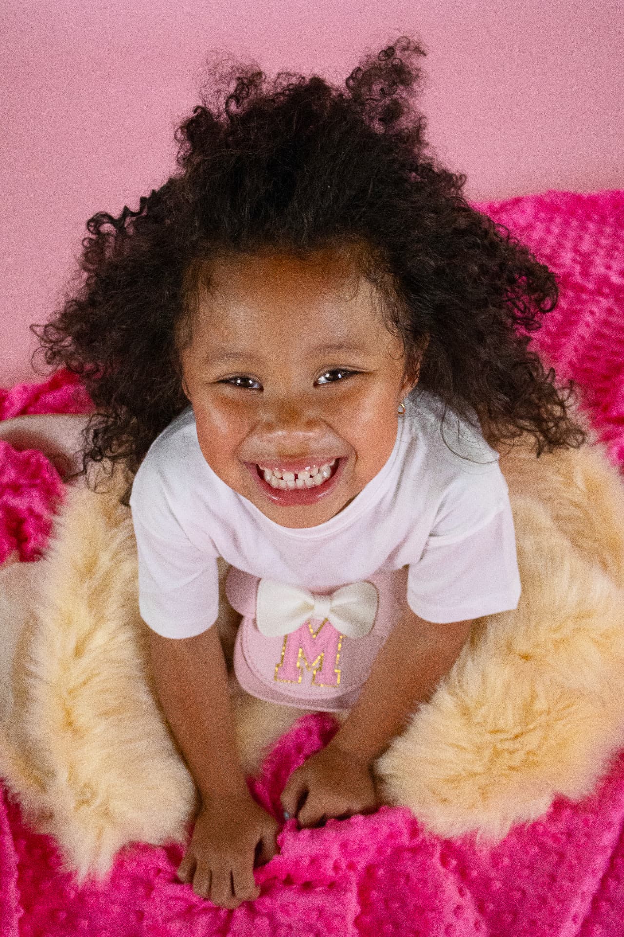 Milani smiling brightly on a pink blanket in a white T-shirt with a bow detail