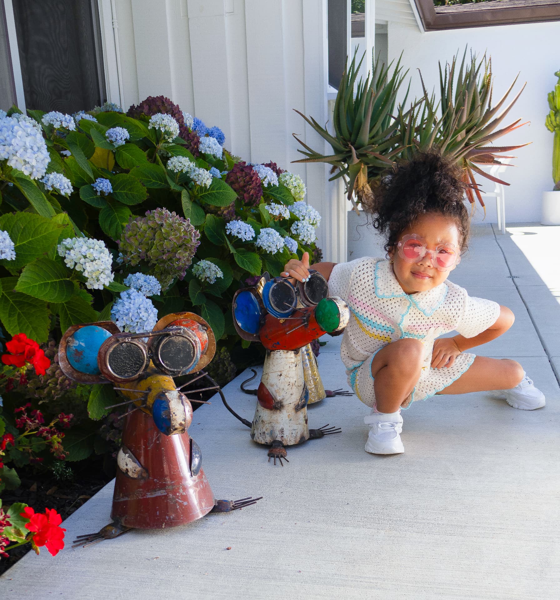 Milani posing outdoors in a crocheted outfit and flower sunglasses beside colorful garden sculptures
